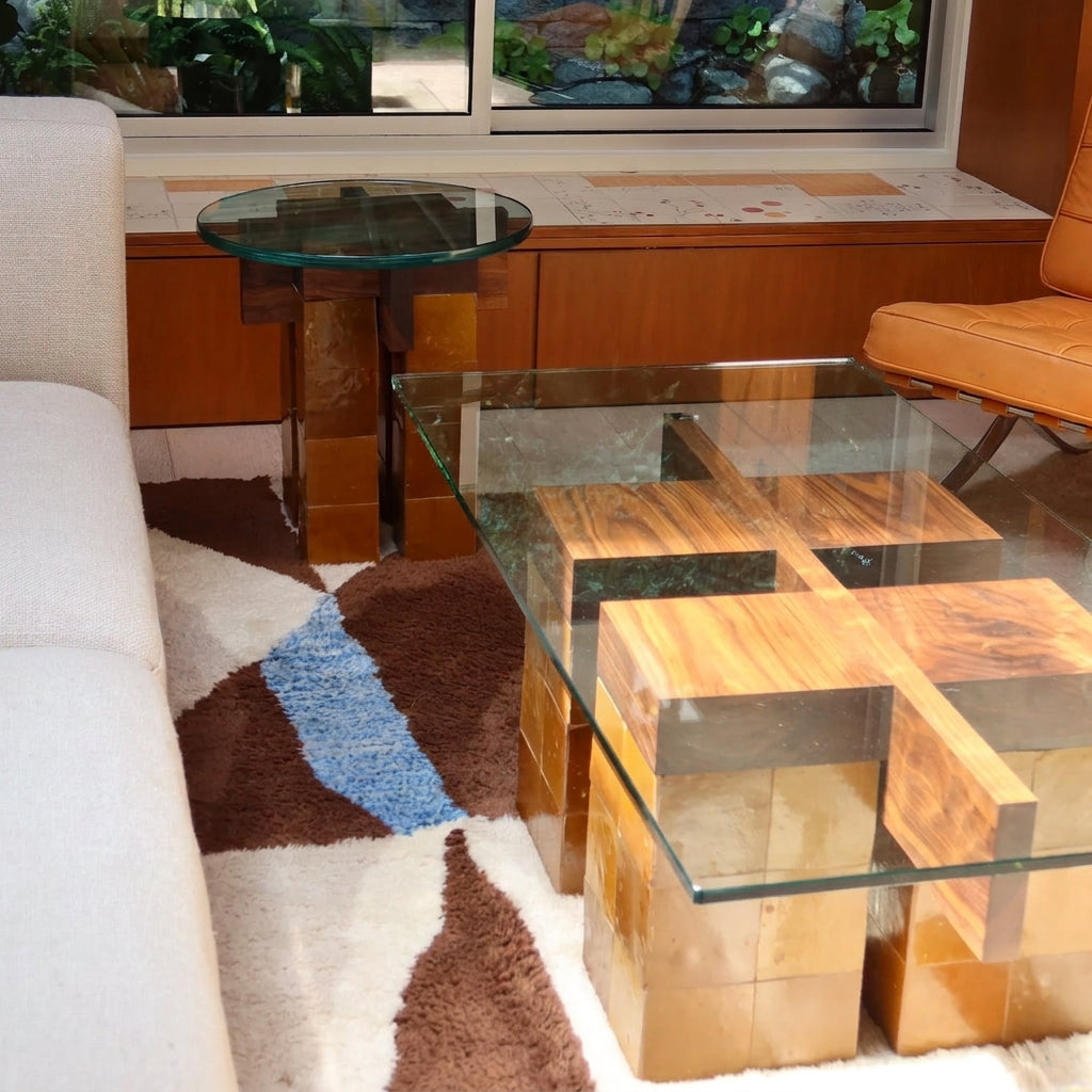 A modern glass-top coffee table with geometric wood block bases, shown in a living room setting next to a white sofa and a patterned rug.