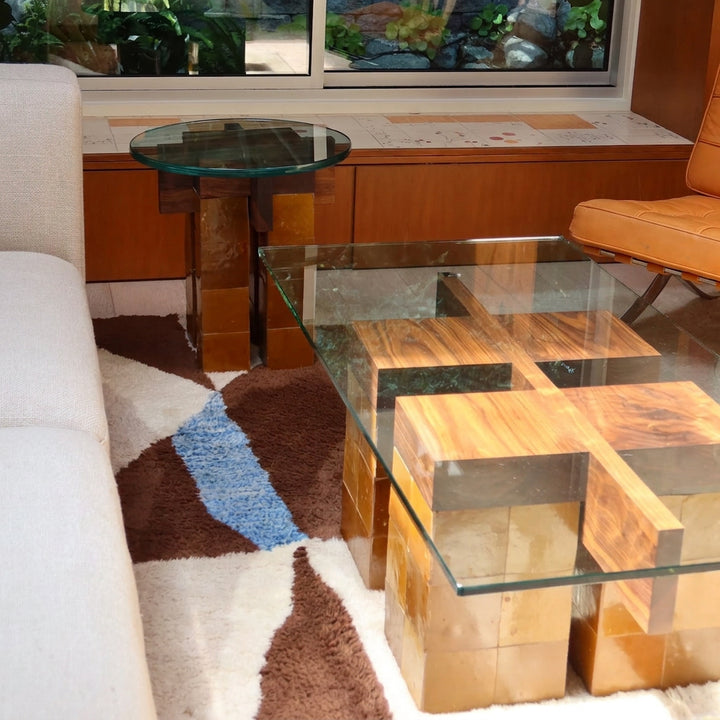 A modern glass-top coffee table with geometric wood block bases, shown in a living room setting next to a white sofa and a patterned rug.