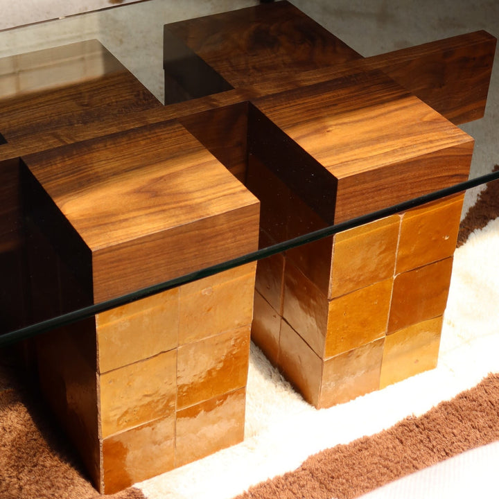 A modern geometric coffee table with a clear glass top and two blocky wooden bases made of stacked wood panels, shown in a room setting on a patterned rug.