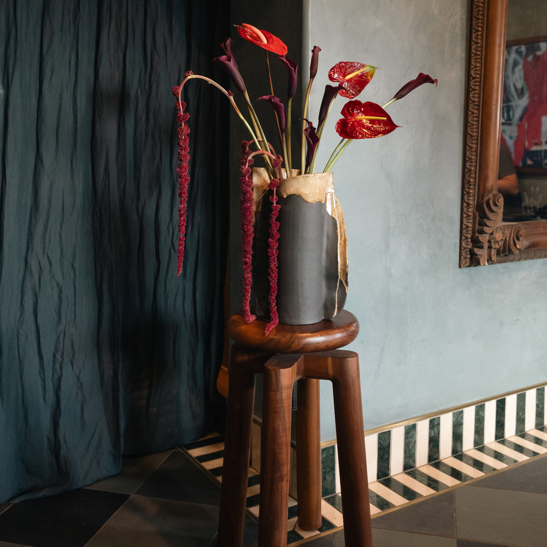 Decorative vase with red flowers on a wooden stool against a textured wall.
