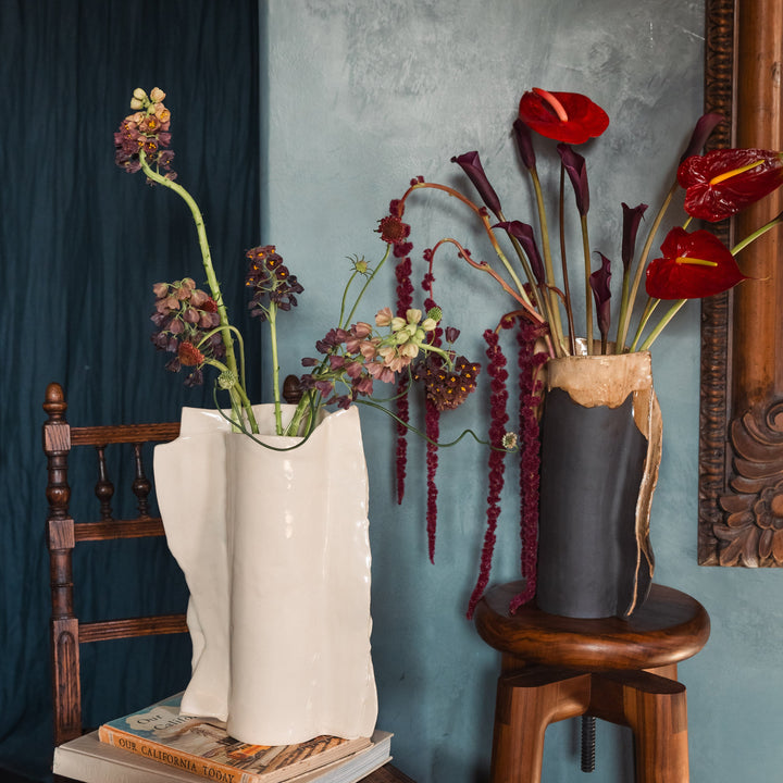 Beige or ivory ceramic vase in high gloss with dried plants and flowers on a wooden vintage chair against a gray lime wash wall with an ornate floral mirror.