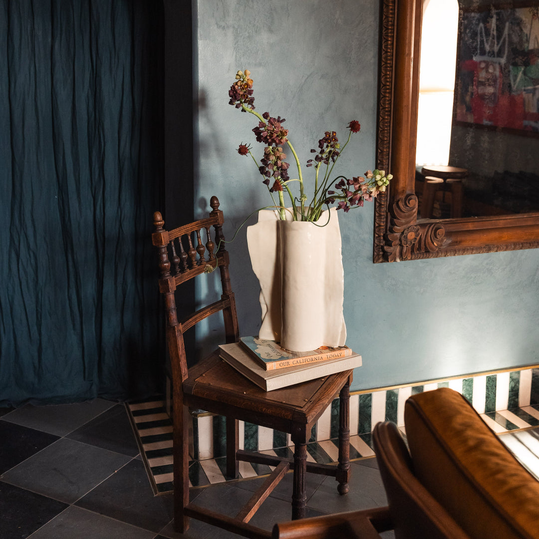Beige or ivory ceramic vase in high gloss with dried plants and flowers on a wooden vintage chair against a gray lime wash wall with an ornate floral mirror.