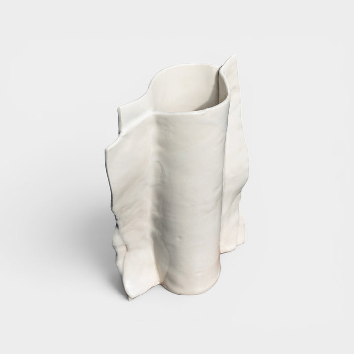 White ceramic vase art form with wavy design on a light gray background Beige or ivory ceramic vase in high gloss with dried plants and flowers on a wooden vintage chair against a gray lime wash wall with an ornate floral mirror.