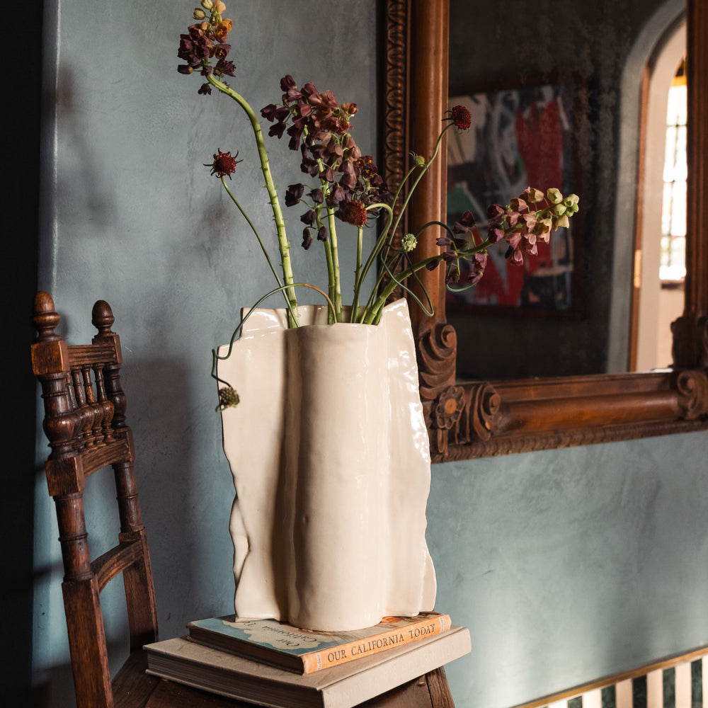Beige or ivory ceramic vase in high gloss with dried plants and flowers on a wooden vintage chair against a gray lime wash wall with an ornate floral mirror.