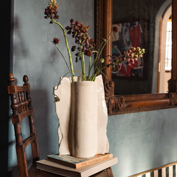 Beige or ivory ceramic vase in high gloss with dried plants and flowers on a wooden vintage chair against a gray lime wash wall with an ornate floral mirror.
