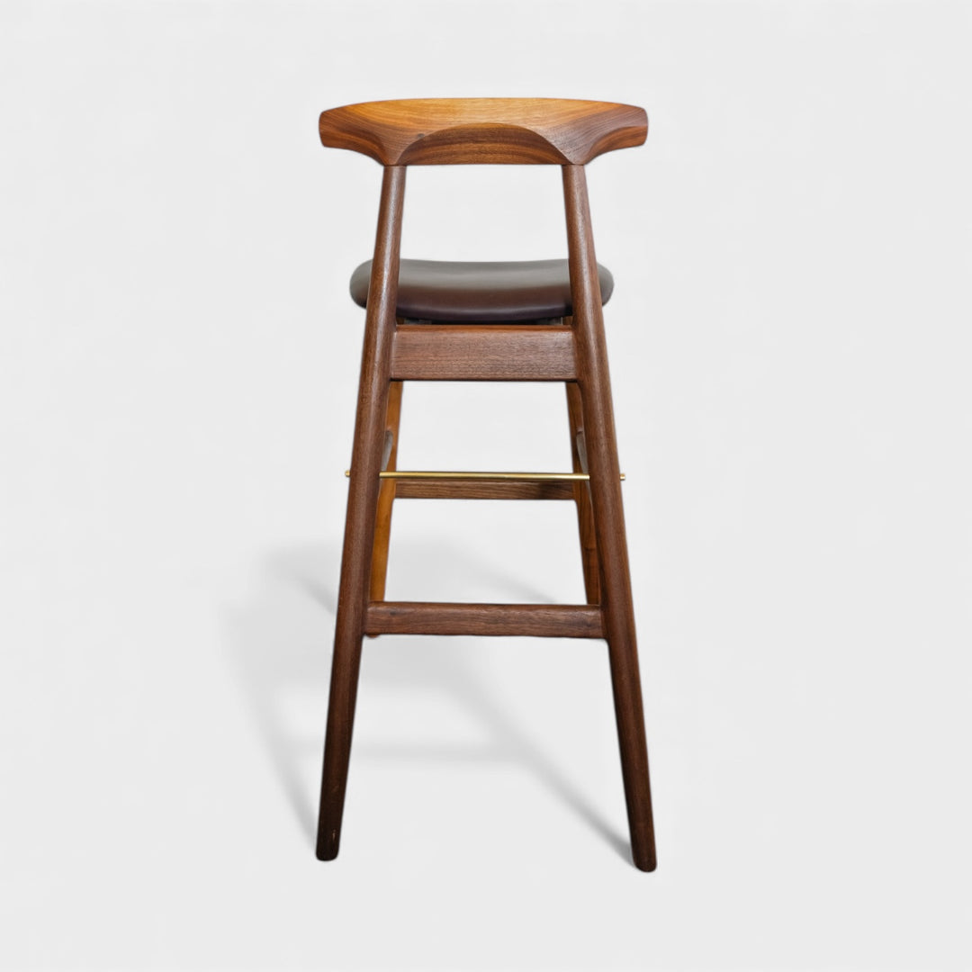 The WOOD High Stool (Bar Height) by Earl Home is a handcrafted wooden bar stool with a light finish, backrest, footrest, and simple design. It offers customizable upholstery to match any decor.