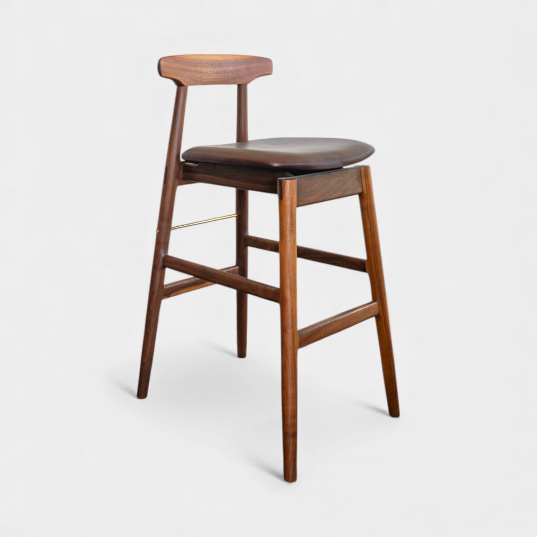 The WOOD High Stool (Bar Height) by Earl Home is a handcrafted wooden bar stool with a light finish, backrest, footrest, and simple design. It offers customizable upholstery to match any decor.