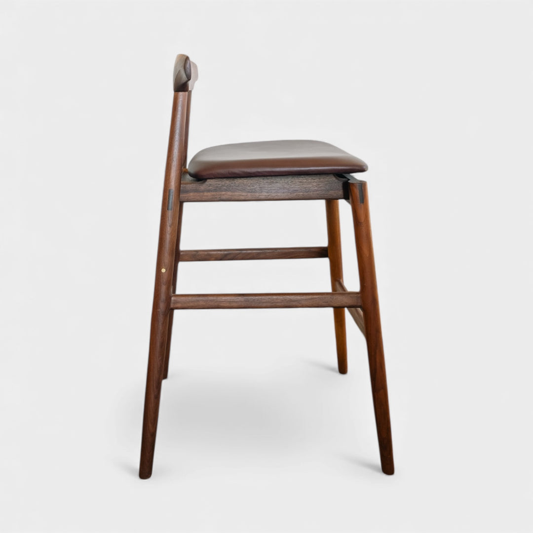 The WOOD High Stool (Bar Height) by Earl Home is a handcrafted wooden bar stool with a light finish, backrest, footrest, and simple design. It offers customizable upholstery to match any decor.