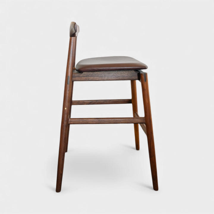 The WOOD High Stool (Bar Height) by Earl Home is a handcrafted wooden bar stool with a light finish, backrest, footrest, and simple design. It offers customizable upholstery to match any decor.