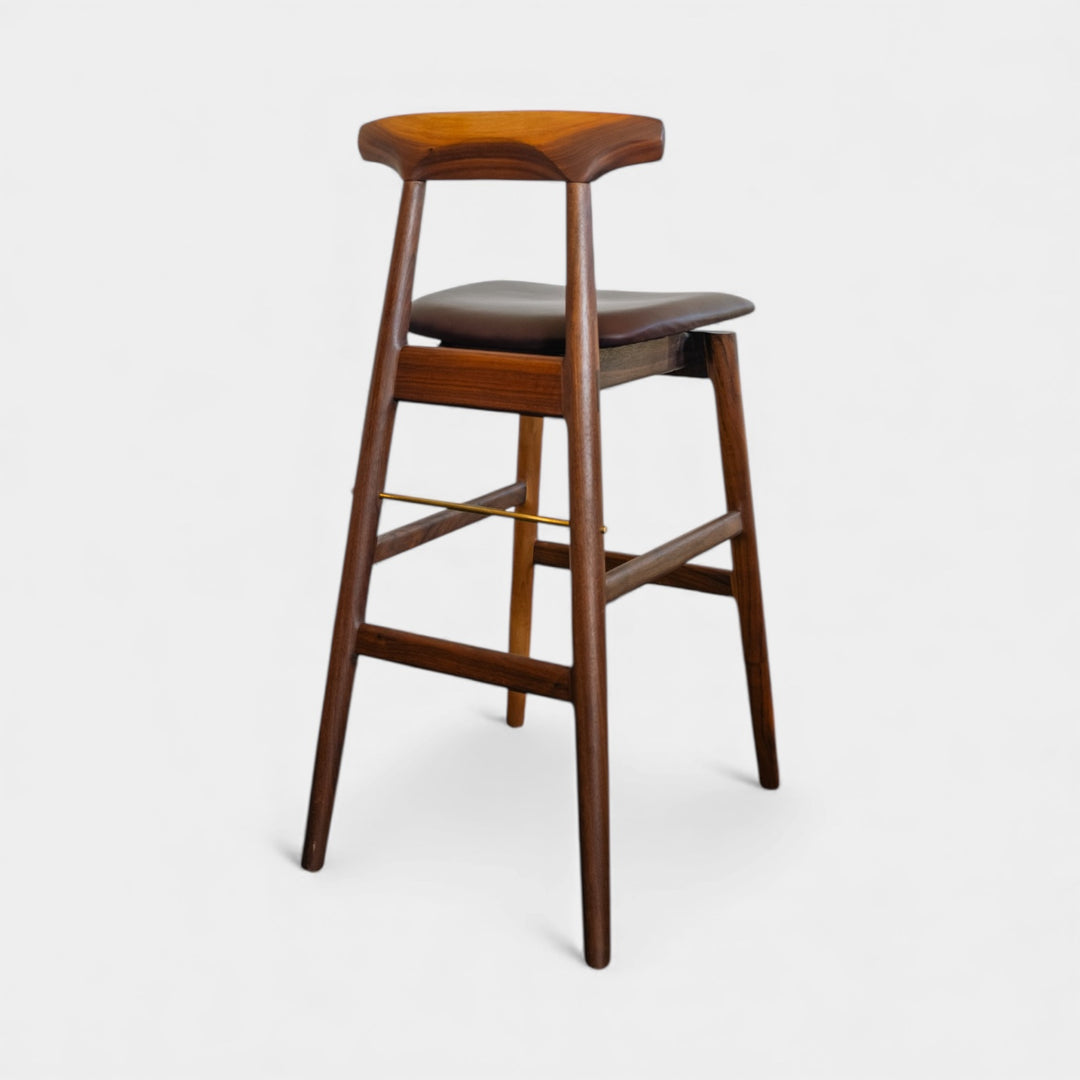The WOOD High Stool (Bar Height) by Earl Home is a handcrafted wooden bar stool with a light finish, backrest, footrest, and simple design. It offers customizable upholstery to match any decor.