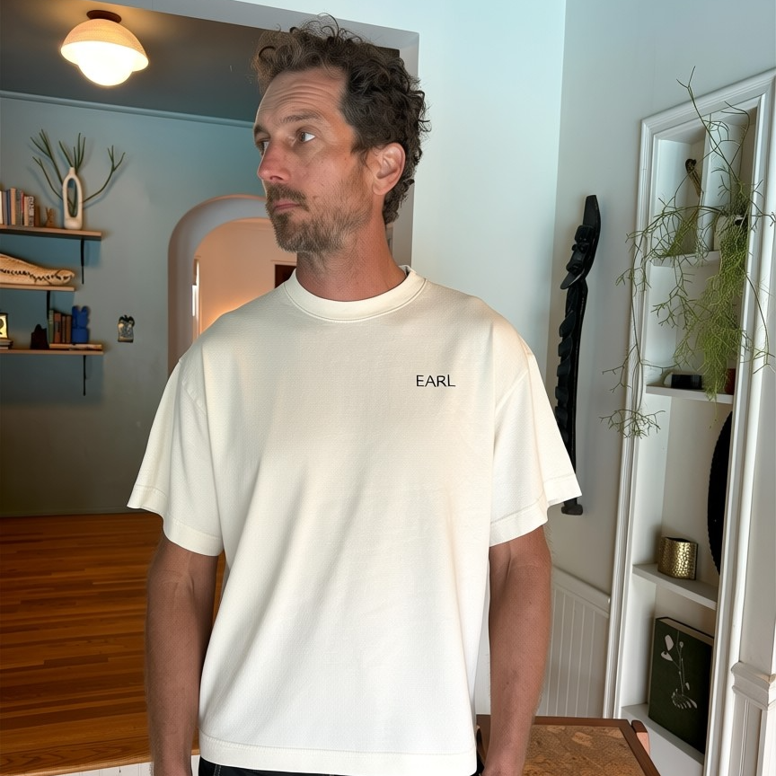 Man wearing a white t-shirt with 'EARL' logo in a room with wooden furniture and shelves.