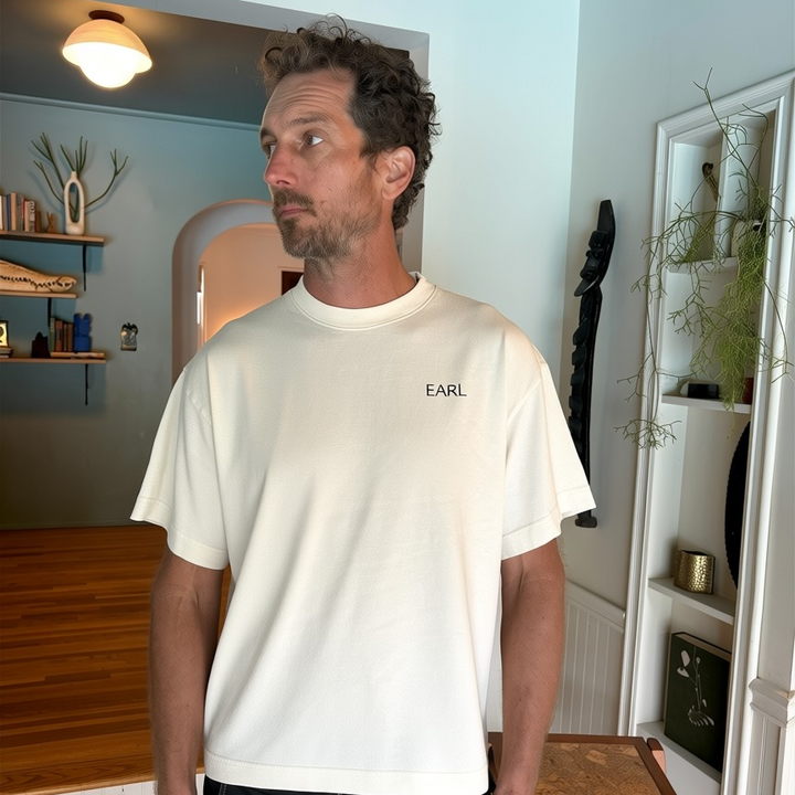 Man wearing a white t-shirt with 'EARL' logo in a room with wooden furniture and shelves.