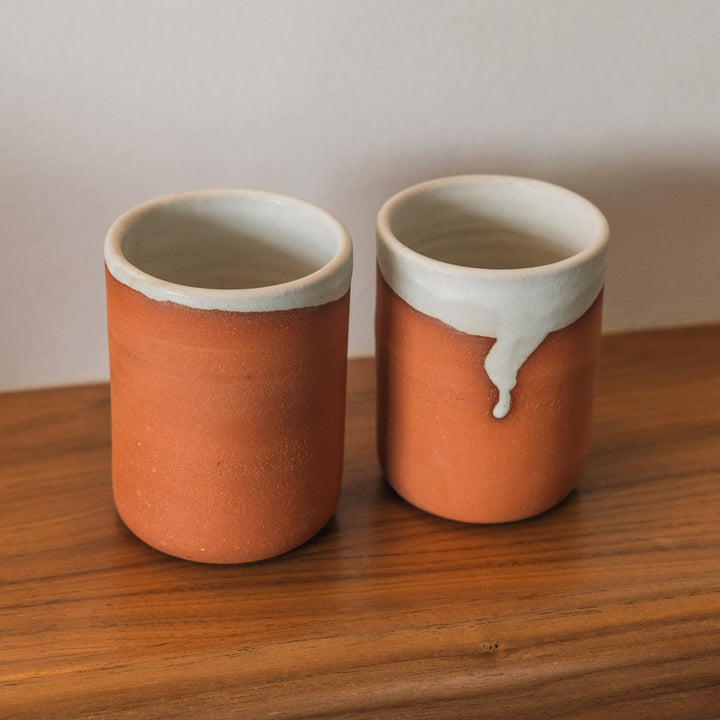 Two ceramic cups with orange-brown bodies and white rims on a wooden surface.