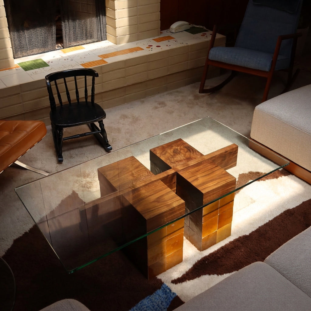 A modern glass-top coffee table with geometric wooden block bases, placed in a living room setting with a fireplace, chair, and sofa visible.