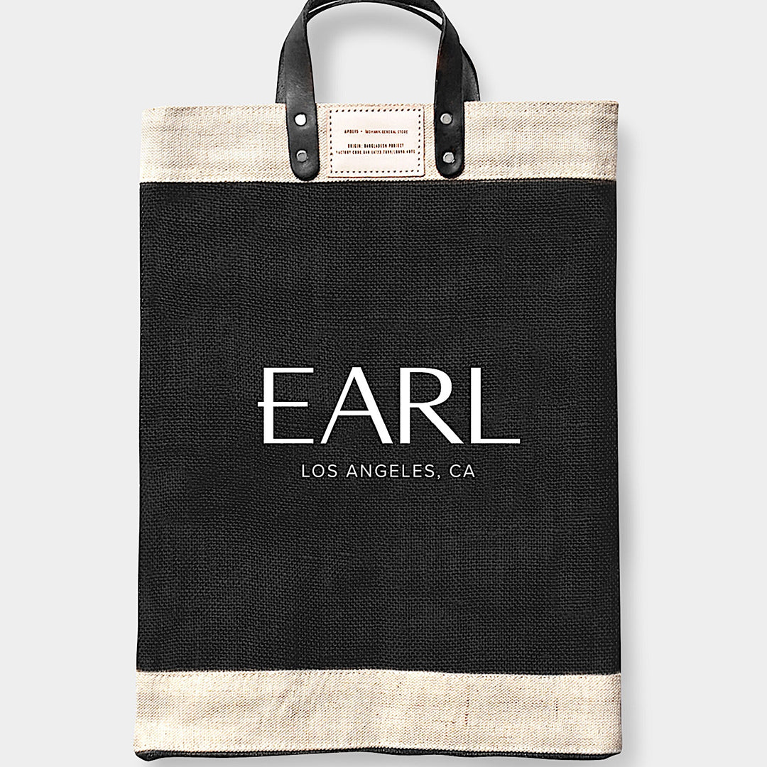 The LARGE EARL * Apolis Market Bag by Earl Home is a handcrafted, Fair Trade-certified black canvas tote with cream accents and leather handles, made in Bangladesh. It features "EARL LOS ANGELES, CA" printed on the front, making it a stylish choice for market trips.