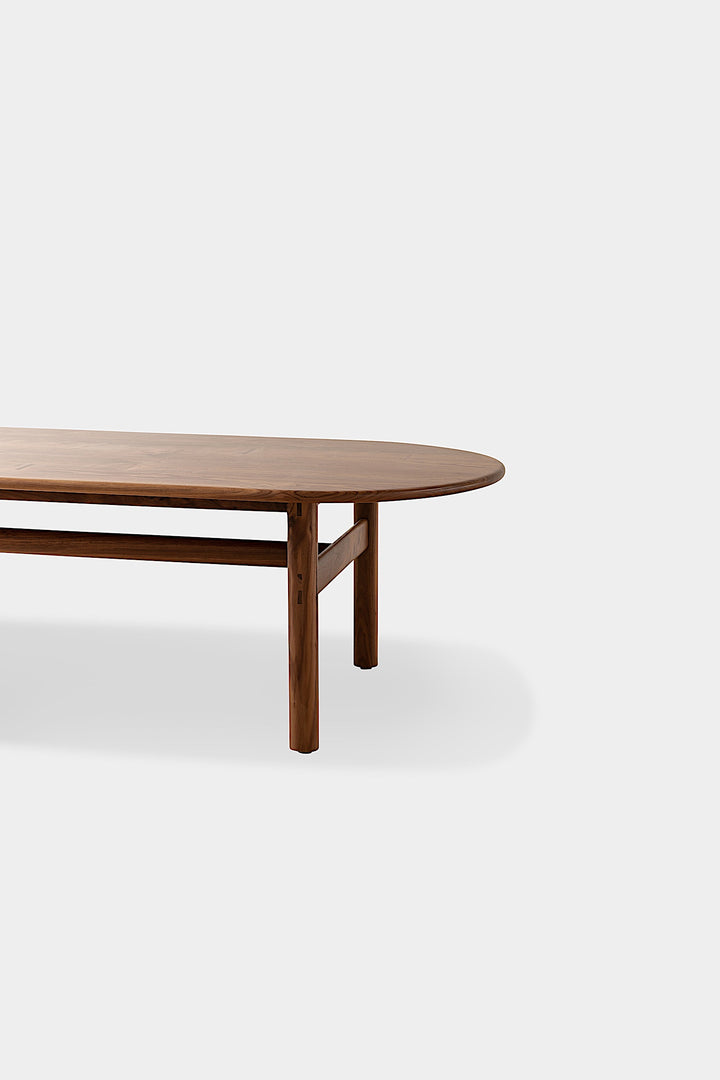 The PALANG Dining Table by Earl Home boasts a robust oval design in rich walnut, supported by four legs and a crossbeam. It comes on a plain white background and offers customizable lengths for perfect fitting in any space.