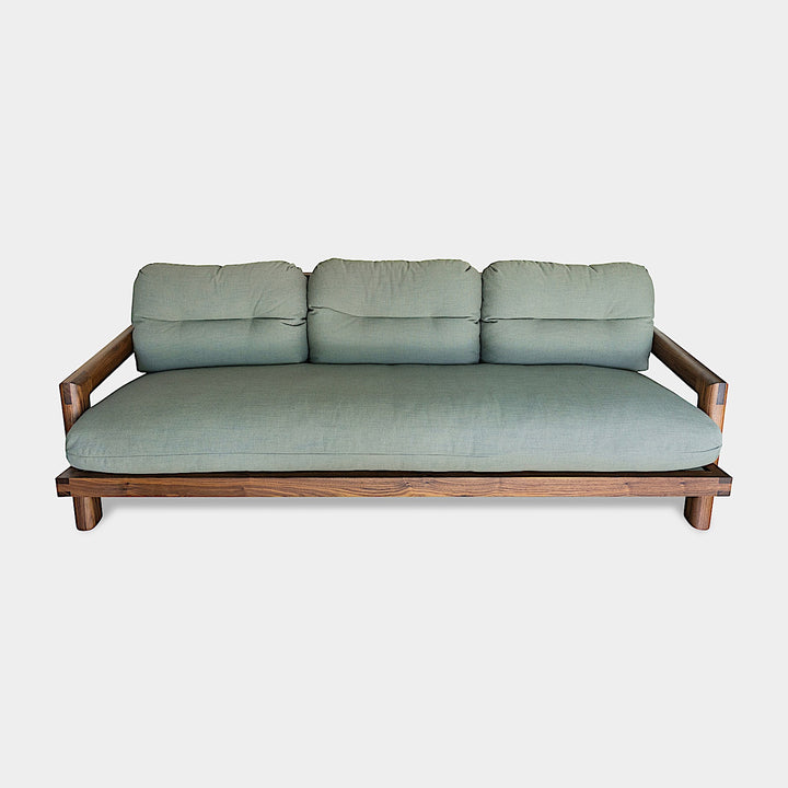 Earl Home's TAMBU Sofa features an American walnut wooden frame with green cushions and three backrests against a minimalist background. Handmade to order, it seamlessly blends elegance with bespoke craftsmanship in its striking design.