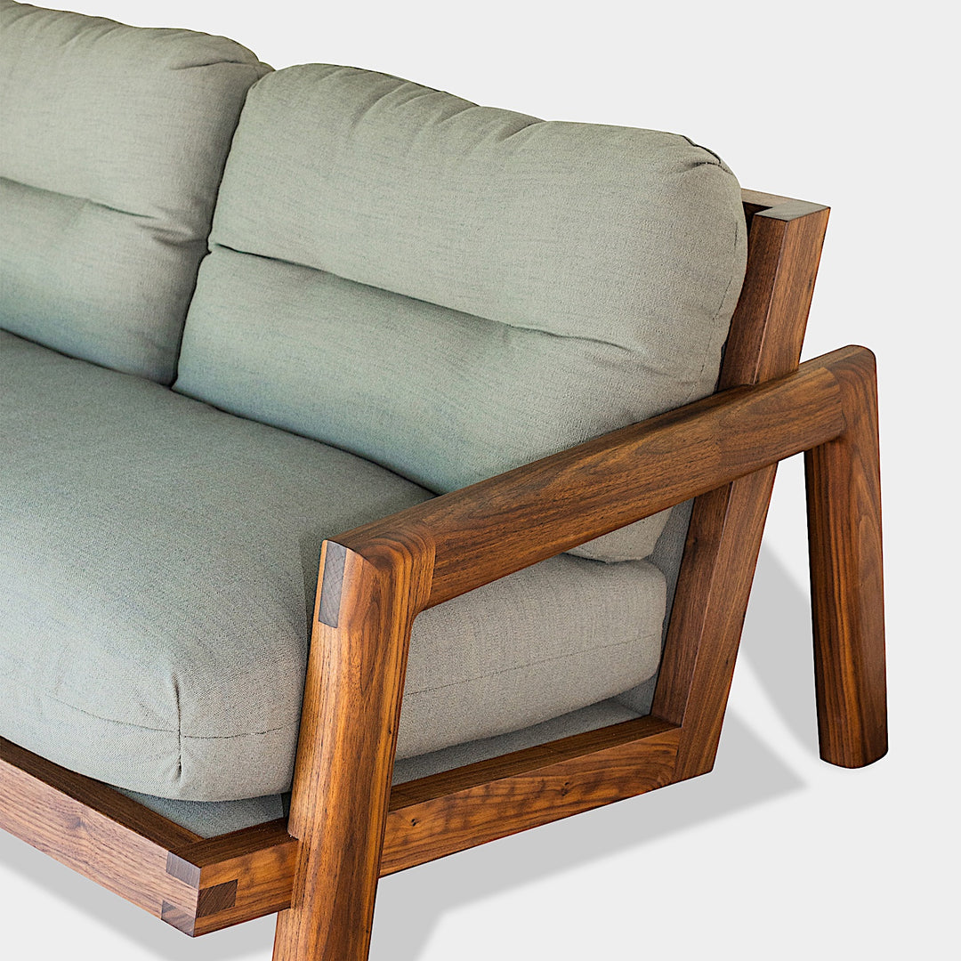 Earl Home's TAMBU Sofa features an American walnut wooden frame with green cushions and three backrests against a minimalist background. Handmade to order, it seamlessly blends elegance with bespoke craftsmanship in its striking design.