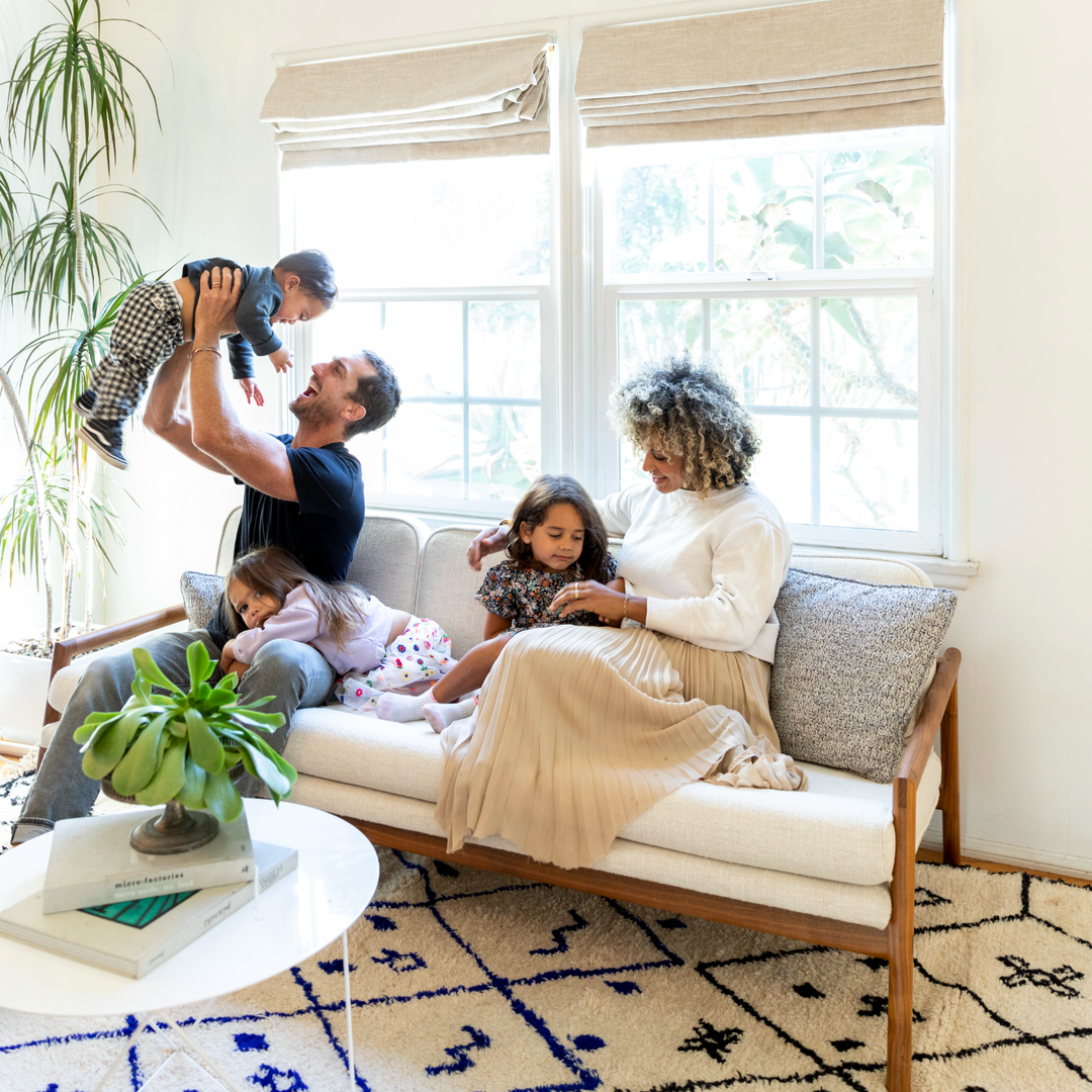 A man and woman sit on a couch with three children, one lifting a baby, in a bright living room with a plant and a round table.