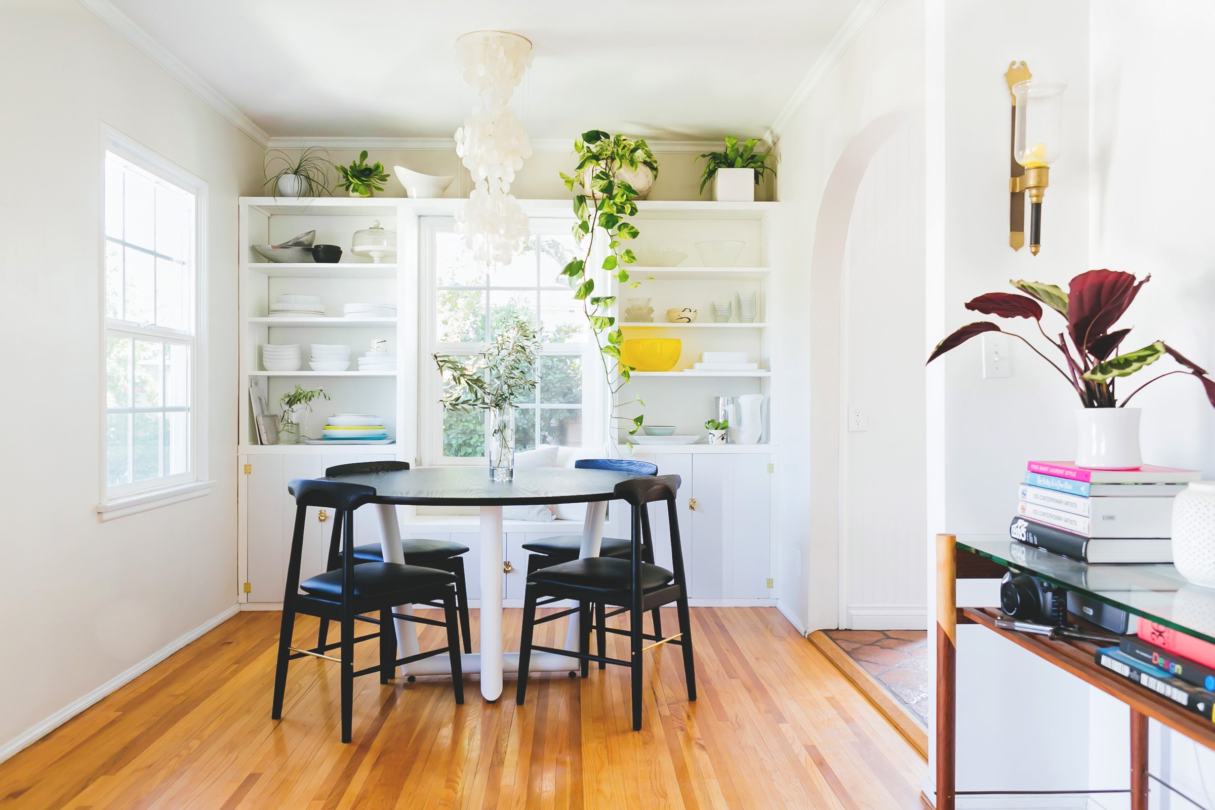 Bright dining room with a round white table, four black chairs, and a wall of shelves displaying plants and dishes. Hardwood floors and large windows provide natural light.