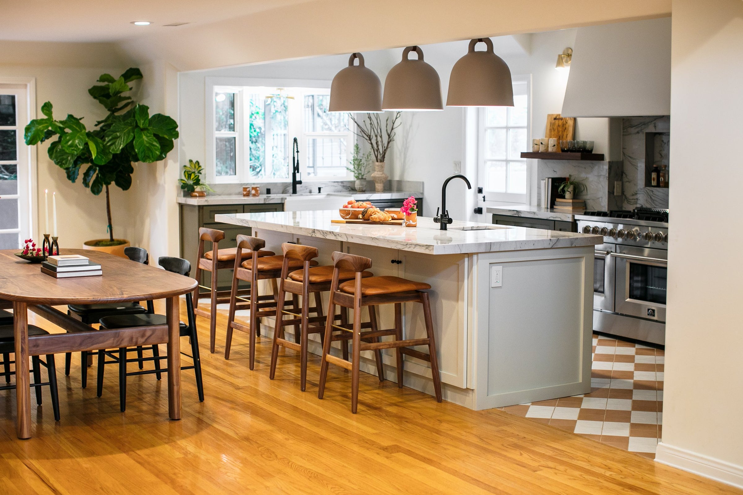 A modern kitchen and dining area with wooden flooring, a large island with stools, pendant lights, a dining table, and a potted plant by the window.