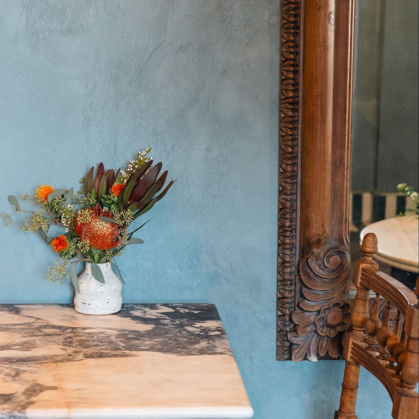 Vintage-style room with wooden cane chair, marble stone table, and decorative ornate floral mirror against a blue limewash portola paint wall.