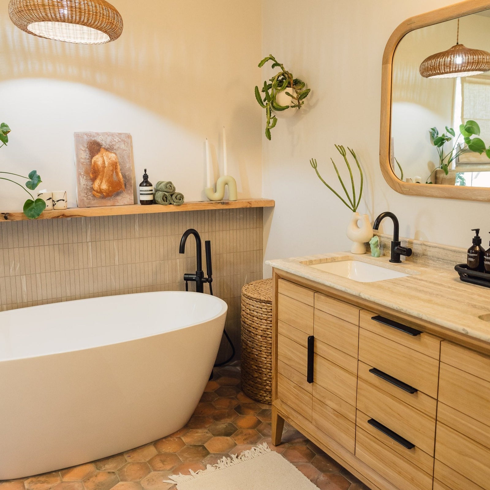 A bathroom with a freestanding tub, wooden vanity, round mirror, and plants. There are candles and a small artwork on a shelf. Brick flooring and natural light enhance the cozy atmosphere.