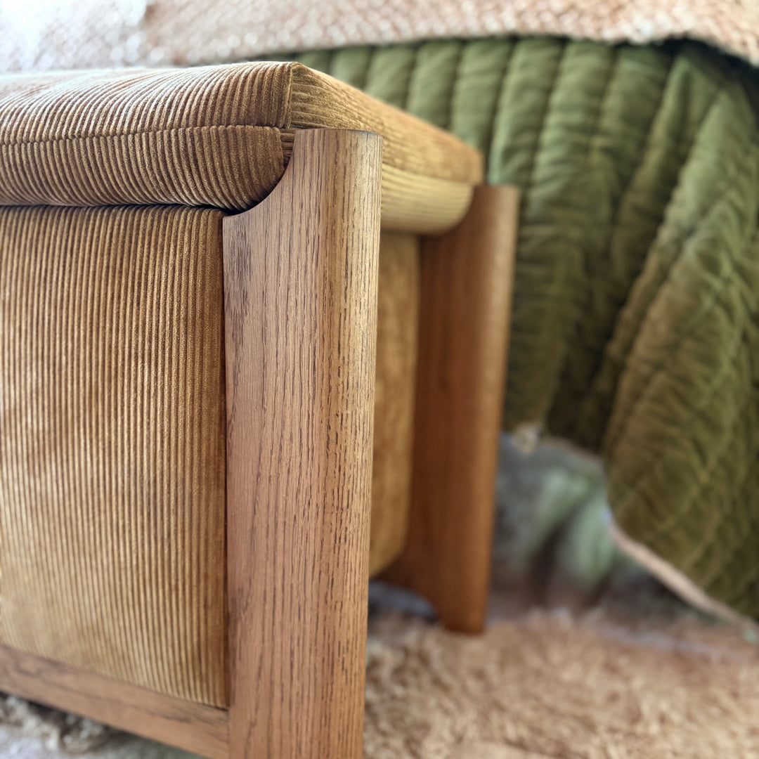 A brown velvet corduroy upholstered storage bench with wooden legs placed at the foot of a bed in a styled bedroom setting.