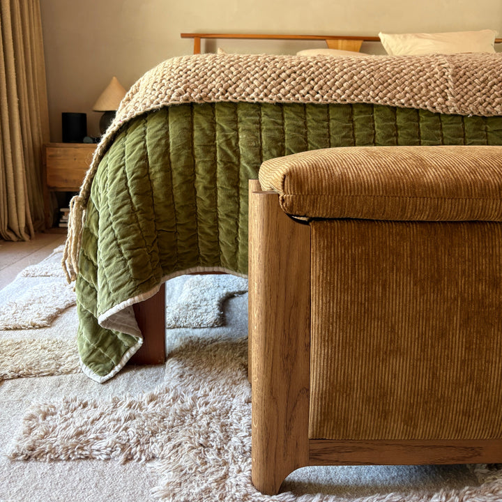 A brown velvet corduroy upholstered storage bench with wooden white oak legs placed at the foot of a bed in a styled bedroom setting.
