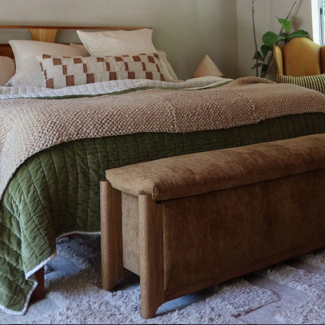 A cozy bedroom interior with a bed dressed in beige and olive green bedding, a brown upholstered bench at the foot of the bed, a wood-paneled ceiling, and a window with greenery outside.