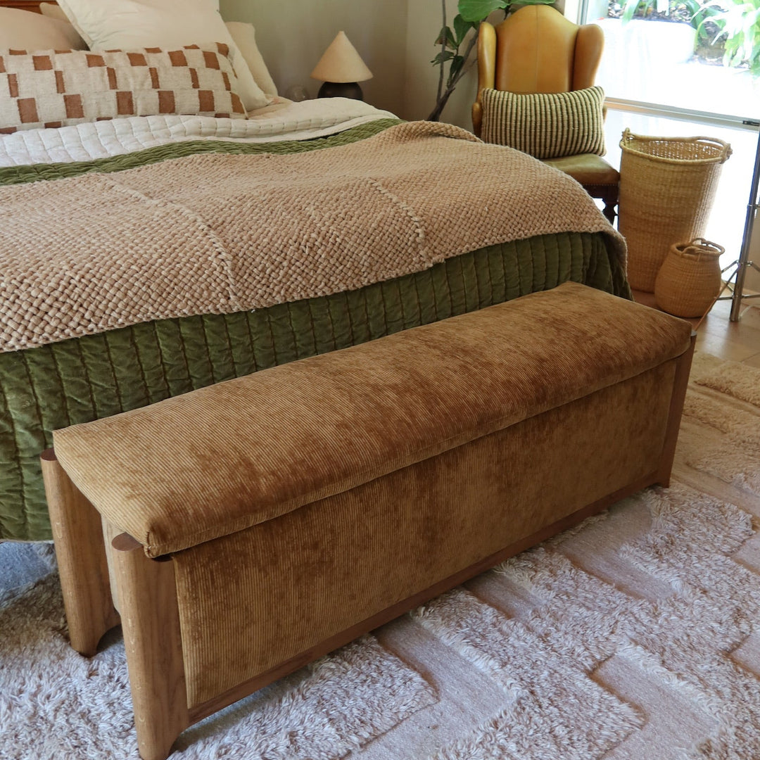 A brown velvet corduroy upholstered storage bench with wooden legs placed at the foot of a bed in a styled bedroom setting.