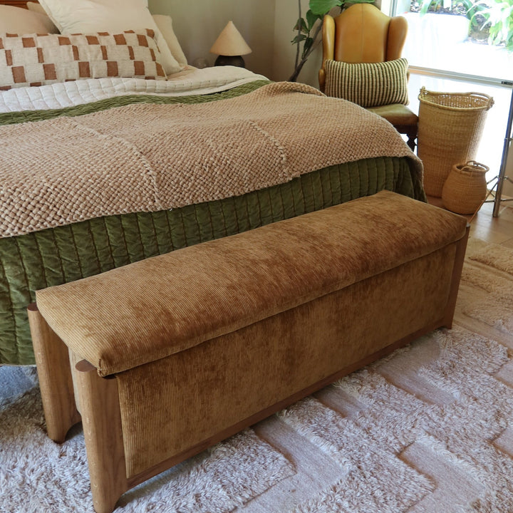 A brown velvet corduroy upholstered storage bench with wooden legs placed at the foot of a bed in a styled bedroom setting.