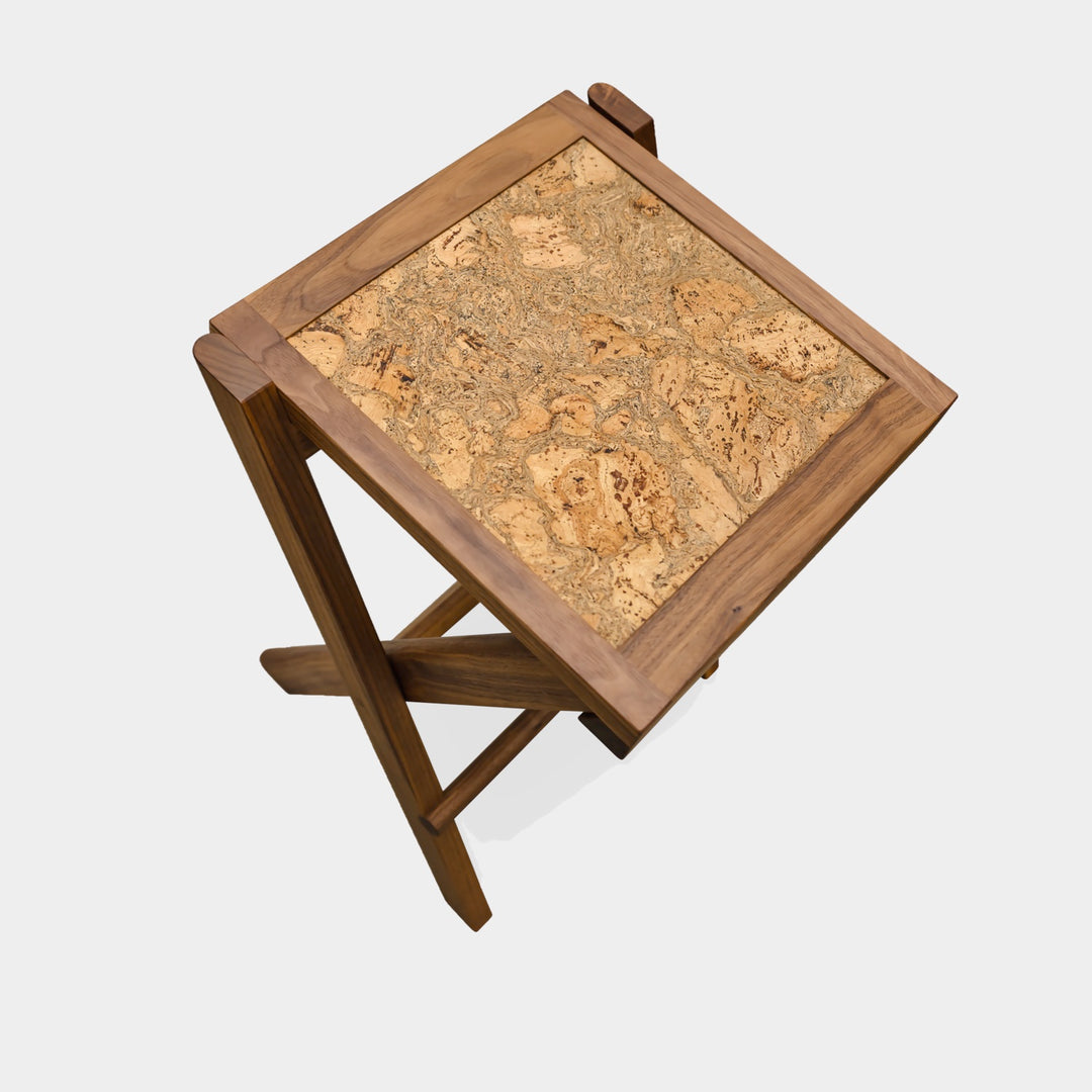 The LAPILO Folding Stool by Earl Home features an angular solid wood frame with a marbled cork seat and cross-legged design in a sleek finish.