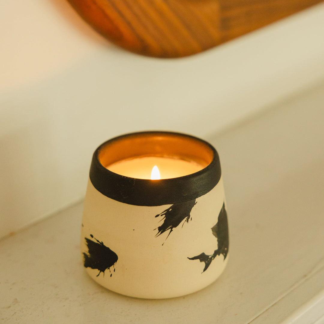 The Earl Home CANDLE is a white ceramic votive with a black splatter pattern, featuring an unlit cardamom-scented candle, set against a gray background.