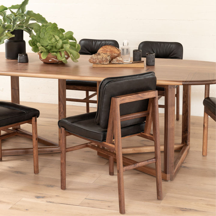 The MORESBY Dining Chair by Earl Home features a custom upholstered black leather seat and backrest, elegantly crafted on a solid wood walnut frame with simple, angular lines.