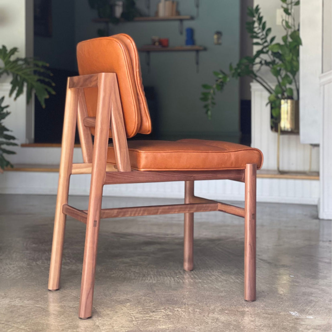 The MORESBY Dining Chair by Earl Home features a custom upholstered camel leather seat and backrest, elegantly crafted on a solid wood walnut frame with simple, angular lines.