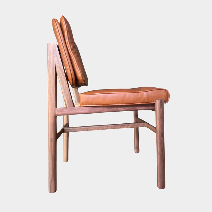 The MORESBY Dining Chair by Earl Home features a custom upholstered camel leather seat and backrest, elegantly crafted on a solid wood walnut frame with simple, angular lines.