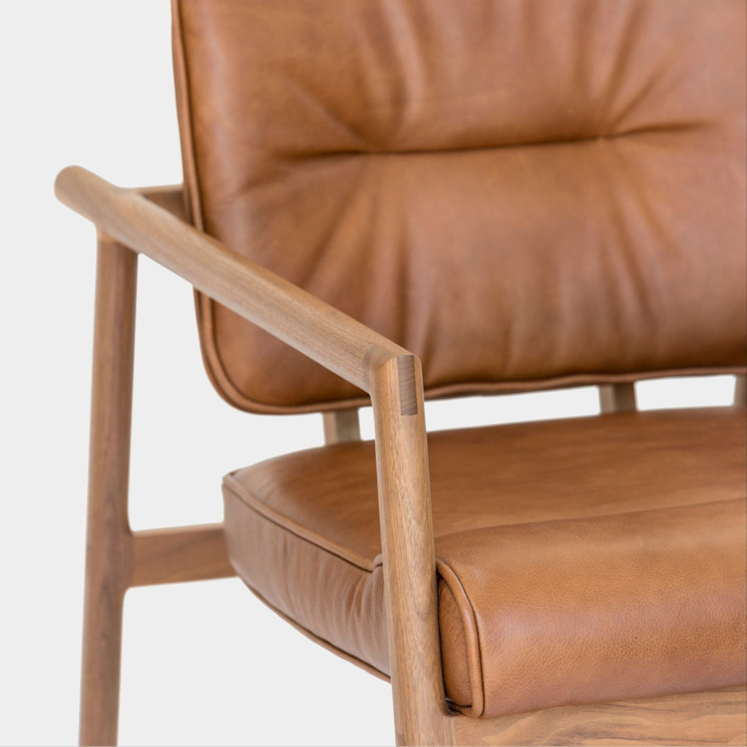 The MORESBY Lounge Chair by Earl Home is a modern, handmade piece featuring a sleek walnut frame and a custom upholstered camel leather cushion.