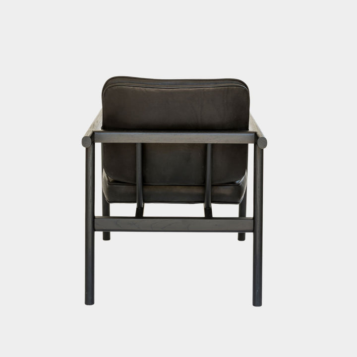 The MORESBY Lounge Chair by Earl Home is a modern, handmade piece featuring a sleek black ebonized oak frame and a custom upholstered black leather cushion.
