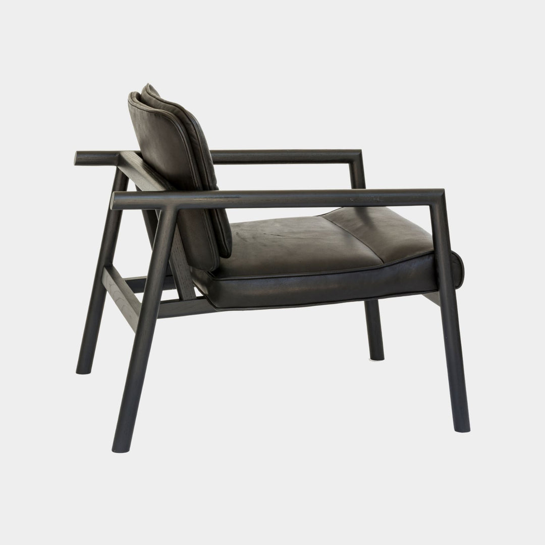 The MORESBY Lounge Chair by Earl Home is a modern, handmade piece featuring a sleek black ebonized oak frame and a custom upholstered black leather cushion.