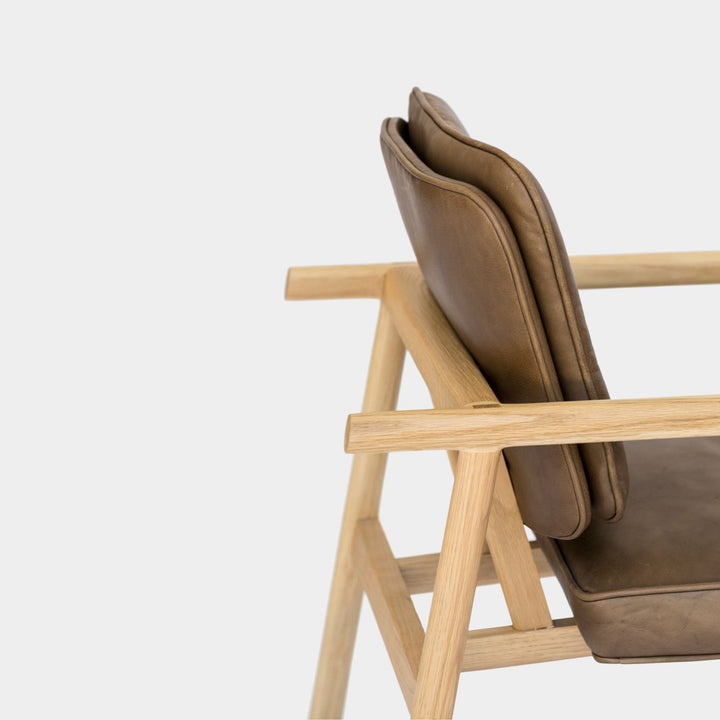 The MORESBY Lounge Chair by Earl Home is a modern, handmade piece featuring a sleek white oak frame and a custom upholstered olive leather cushion.