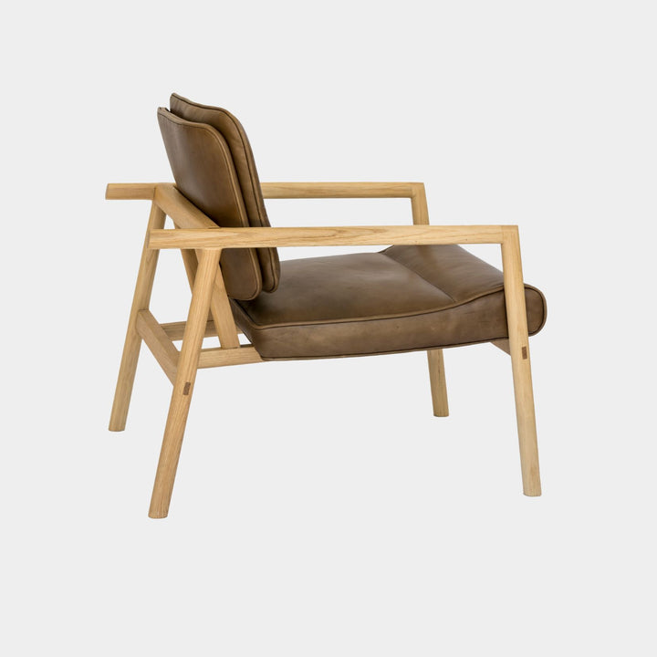 The MORESBY Lounge Chair by Earl Home is a modern, handmade piece featuring a sleek white oak frame and a custom upholstered olive leather cushion.