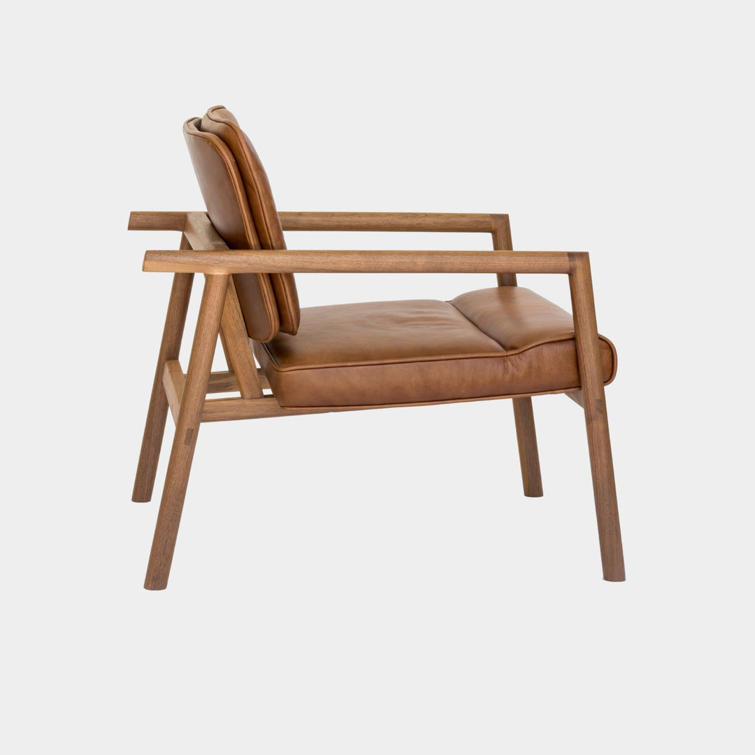 The MORESBY Lounge Chair by Earl Home is a modern, handmade piece featuring a sleek walnut frame and a custom upholstered camel leather cushion.