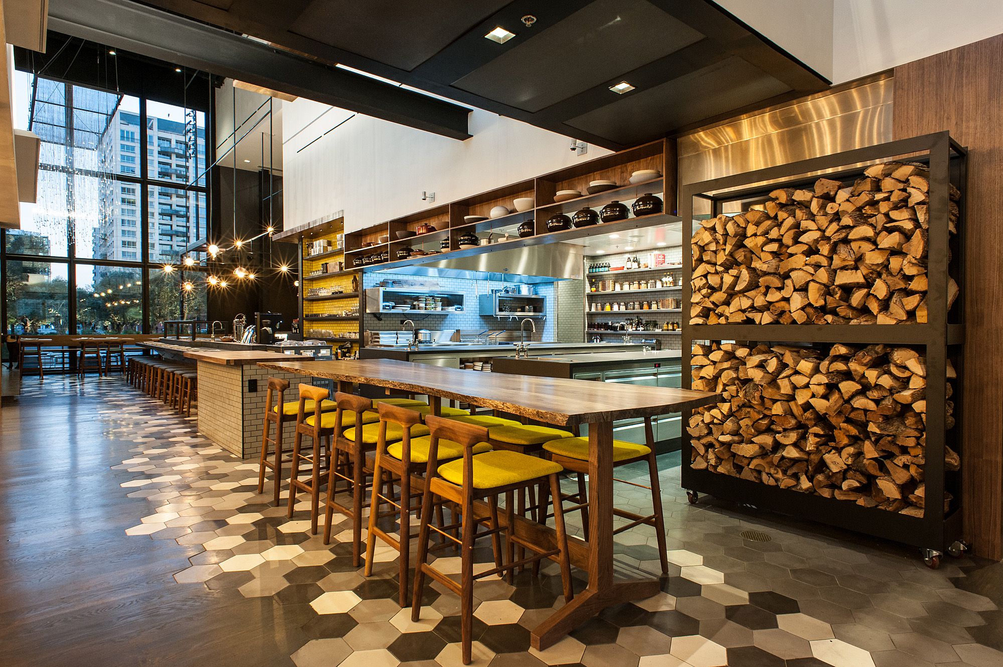 Modern restaurant interior with wooden tables, yellow-cushioned chairs, hexagonal patterned floor, and a wall rack filled with stacked firewood. Large windows display a cityscape outside.