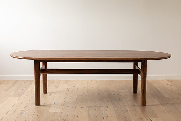 The PALANG Dining Table by Earl Home boasts a robust oval design in rich walnut, supported by four legs and a crossbeam. It comes on a plain white background and offers customizable lengths for perfect fitting in any space.