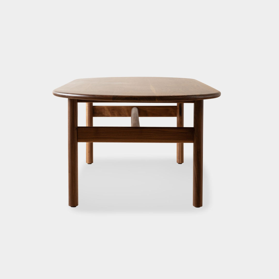 The PALANG Dining Table by Earl Home boasts a robust oval pill shape with bullnose edge design in rich walnut, supported by four legs and a crossbeam. It is available in white oak, maple, or ebonized oak.  This table comes in customizable lengths and top shapes including rectangular or live edge for perfect fitting in any space.