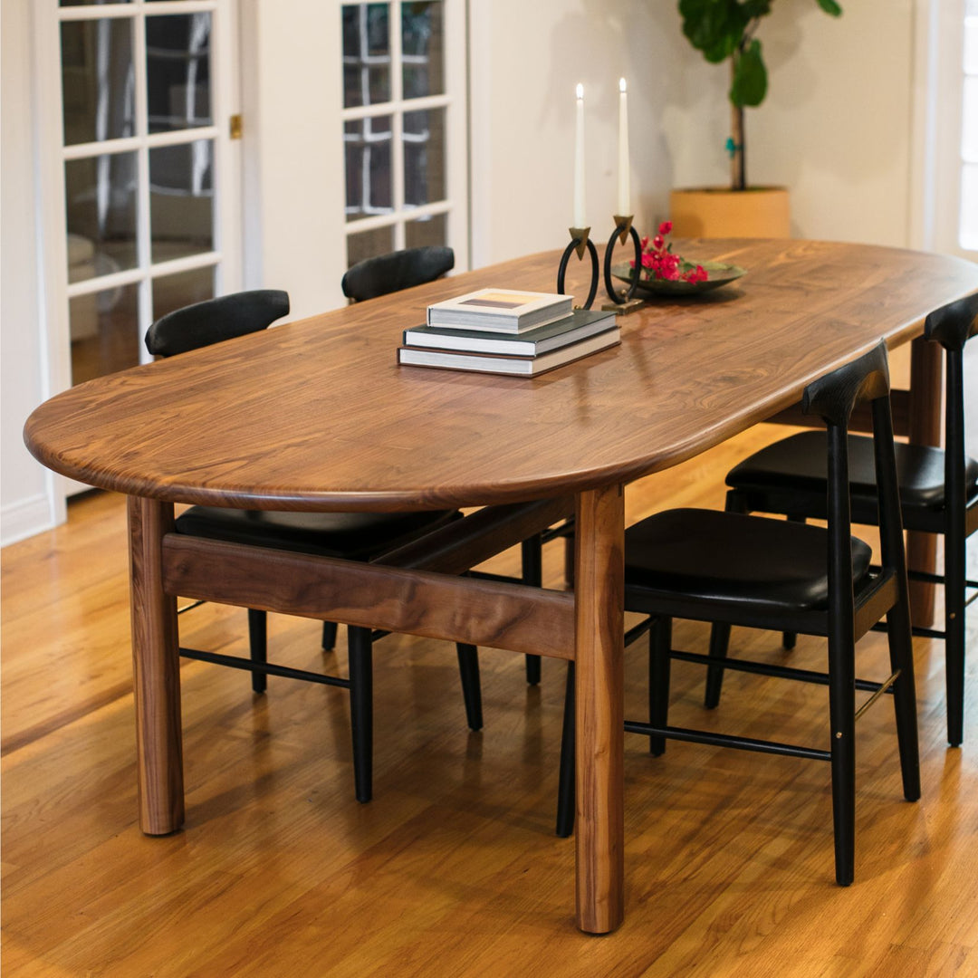 The PALANG Dining Table by Earl Home boasts a robust oval pill shape with bullnose edge design in rich walnut, supported by four legs and a crossbeam. It is available in white oak, maple, or ebonized oak.  This table comes in customizable lengths and top shapes including rectangular or live edge for perfect fitting in any space.