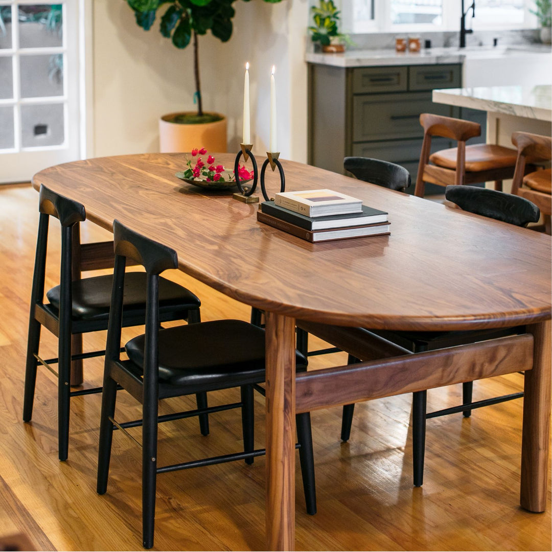 The PALANG Dining Table by Earl Home boasts a robust oval pill shape with bullnose edge design in rich walnut, supported by four legs and a crossbeam. It is available in white oak, maple, or ebonized oak.  This table comes in customizable lengths and top shapes including rectangular or live edge for perfect fitting in any space.