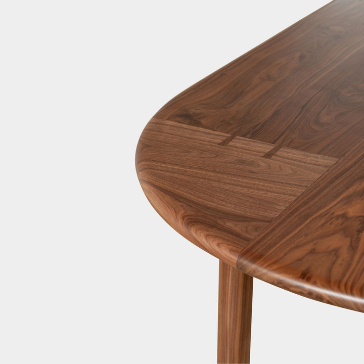 The PALANG Dining Table by Earl Home boasts a robust oval pill shape with bullnose edge design in rich walnut, supported by four legs and a crossbeam. It is available in white oak, maple, or ebonized oak.  This table comes in customizable lengths and top shapes including rectangular or live edge for perfect fitting in any space.