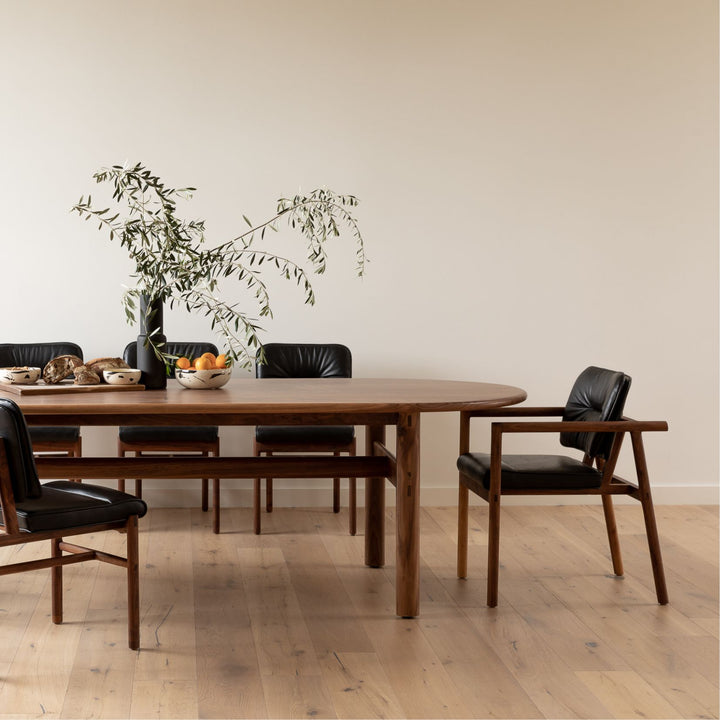 The PALANG Dining Table by Earl Home boasts a robust oval pill shape with bullnose edge design in rich walnut, supported by four legs and a crossbeam. It is available in white oak, maple, or ebonized oak.  This table comes in customizable lengths and top shapes including rectangular or live edge for perfect fitting in any space.